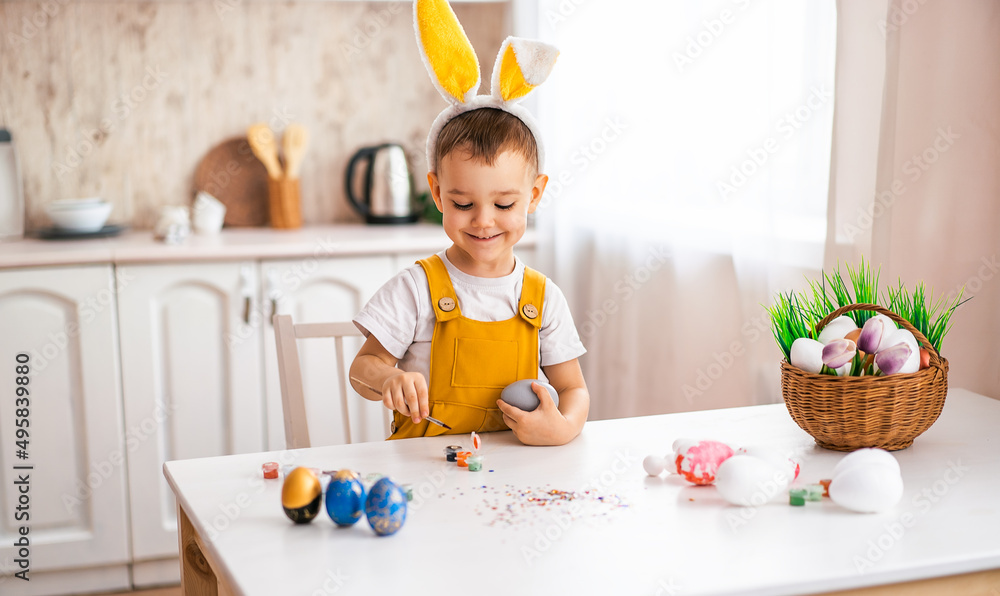 a handsome little boy of European appearance draws on Easter eggs at the table, on his head with yellow rabbit ears. a handsome little boy of European appearance draws on Easter eggs at the table, on his head with yellow rabbit ears.
