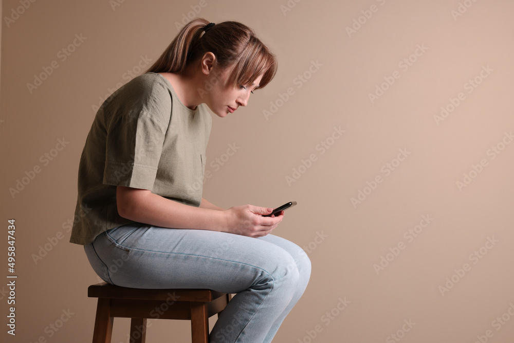 Young woman with poor posture using smartphone while sitting on stool against beige background, space for text - obrazy, fototapety, plakaty 