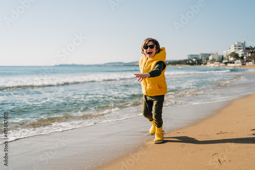 Boy in yellow rubber boots playing with sand at the beach. School kid touching water at autumn winter sea. Child having fun and running. on waves at the shore. Spring Holiday vacation concept.