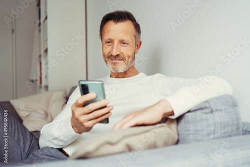 older man sitting at home on sofa and looking at his cell phone photo
