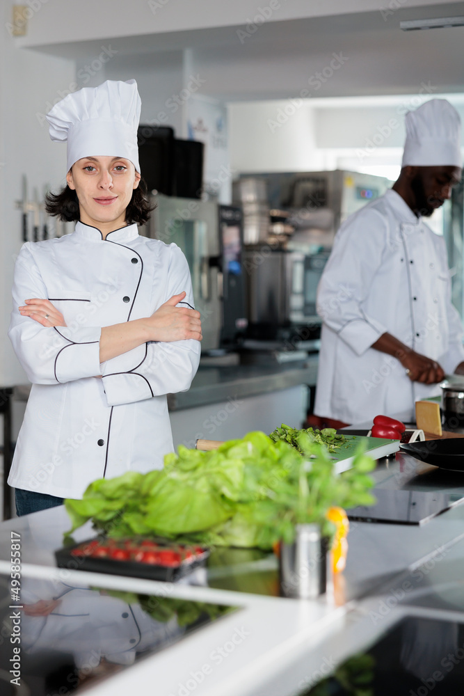 Food industry worker standing in restaurant kitchen with arms crossed after vegetable cutting. Sous chef wearing cooking uniform while preparing ingredients for evening dining service.