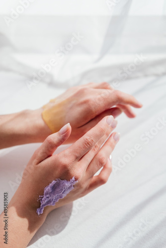Female hands applying purple cream scrub on skin top view white background. Skincare routine concept