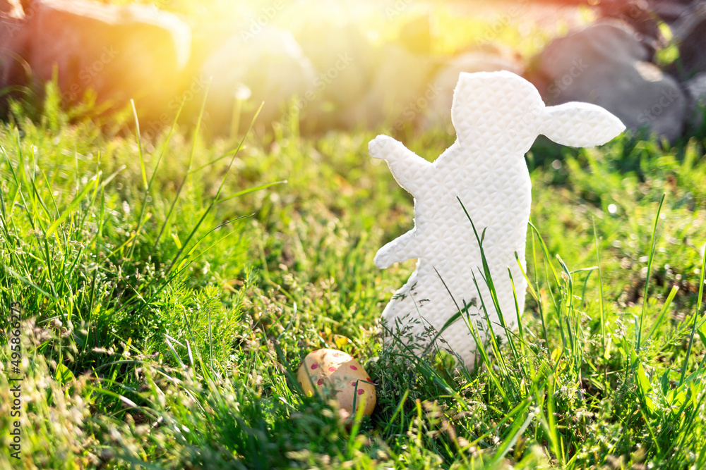 Obraz premium The concept of Easter holidays. Decorative handmade craft in the form of a rabbit, located next to the painted egg. Green grass and stones in the background. Copy space. Light