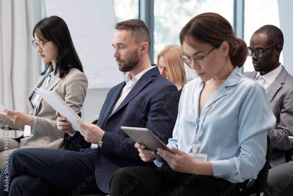 Fototapeta premium Group of intercultural brokers in formalwear looking through paper and electronic documents while taking part in conference