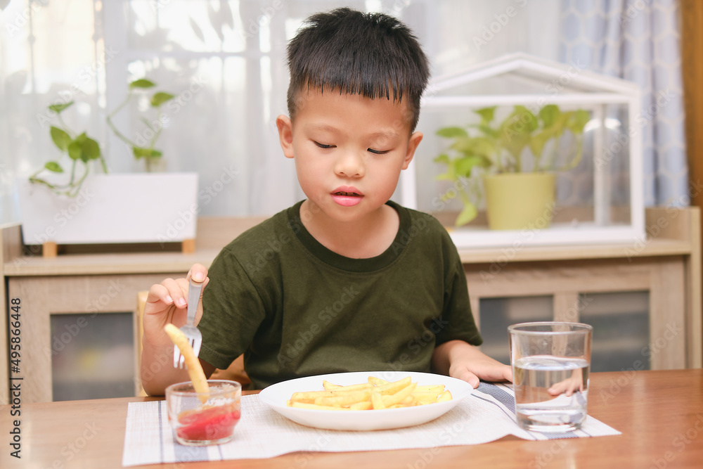 Cute happy smiling Asian 5 yers old kindergarten boy child using fork eating French fries potato chips with ketchup, sitting at the table at home or restaurant, Unhealthy Kids Foods concept Cute happy smiling Asian 5 yers old kindergarten boy child using fork eating French fries potato chips with ketchup, sitting at the table at home or restaurant, Unhealthy Kids Foods concept