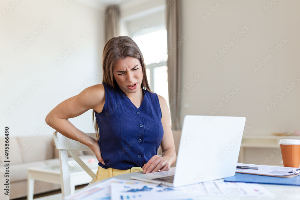 Portrait of young stressed woman sitting at home office desk in front of laptop, touching aching back with pained expression, suffering from backache after working on laptop Portrait of young stressed woman sitting at home office desk in front of laptop, touching aching back with pained expression, suffering from backache after working on laptop