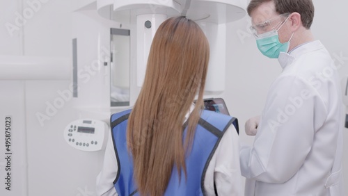On a panoramic x-ray machine, the dentist aligns the bite of a woman. In a dental clinic, a woman dressed in a lead apron stands still as a panoramic X-ray machine circles her head..