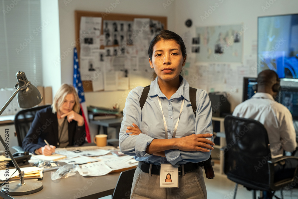 Young Hispanic female leader of intelligence service or police department in uniform crossing arms on chest while standing against coworkers Young Hispanic female leader of intelligence service or police department in uniform crossing arms on chest while standing against coworkers