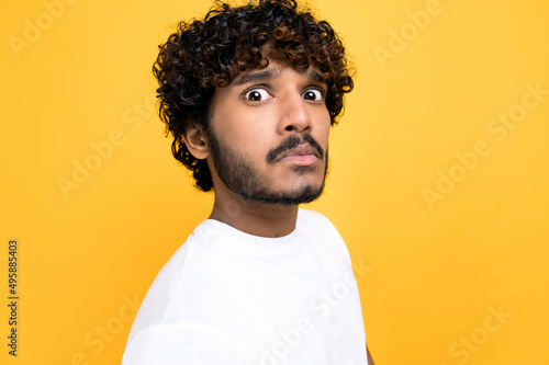 Close-up photo of a scared confused indian or arabian curly haired guy, looking at camera in disbelief, standing over isolated orange background wearing white t-shirt © Kateryna