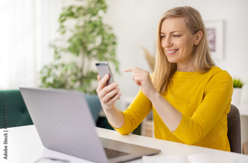 Smiling caucasian woman in glasses sitting at home and using modern smartphone with laptop. Beautiful female blonde having video conversation. Smiling caucasian woman in glasses sitting at home and using modern smartphone with laptop. Beautiful female blonde having video conversation.