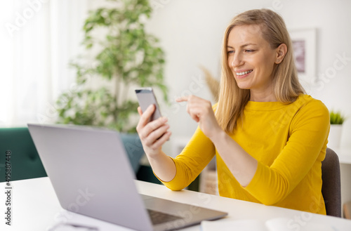 Smiling caucasian woman in glasses sitting at home and using modern smartphone with laptop. Beautiful female blonde having video conversation.