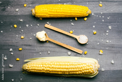 Two cobs of fresh and cooked sweet corn lying on a gray table surrounded by salt and butter on a wooden spoon.
