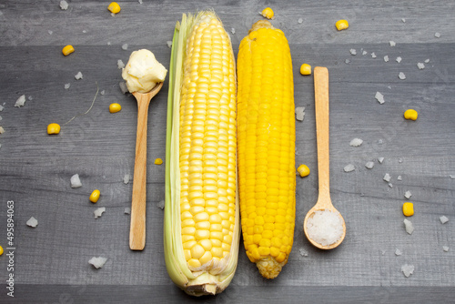 Two cobs of fresh and cooked sweet corn lying on a gray table surrounded by salt and butter on a wooden spoon.
