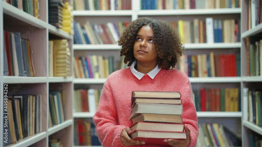 Young Black female wearing pink sweater carrying pile of books in library. Tracking shot student in college library. Concept of education Young Black female wearing pink sweater carrying pile of books in library. Tracking shot student in college library. Concept of education