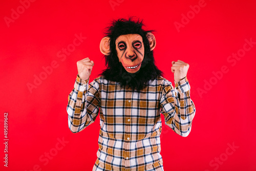 Man with chimpanzee monkey mask and plaid shirt clenching his fists, on red background.