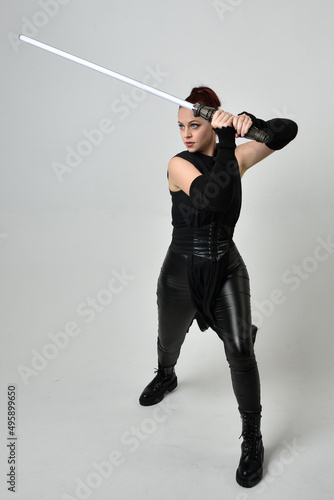 Full length portrait of pretty red haired female model wearing black futuristic scifi leather costume, holding a lightsaber sword weapon. Dynamic standing poses on a white studio background.