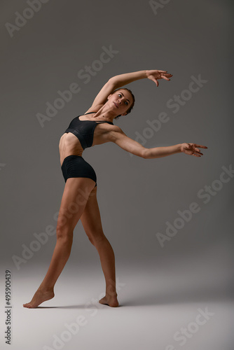 Beautiful gymnast or ballerina in black leotard dancing on a gray studio background