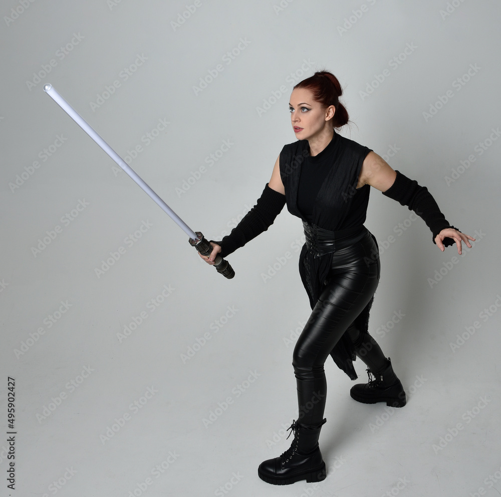 Full length portrait of pretty red haired female model wearing black futuristic scifi leather costume, holding a lightsaber sword weapon. Dynamic standing poses on a white studio background. Full length portrait of pretty red haired female model wearing black futuristic scifi leather costume, holding a lightsaber sword weapon. Dynamic standing poses on a white studio background.