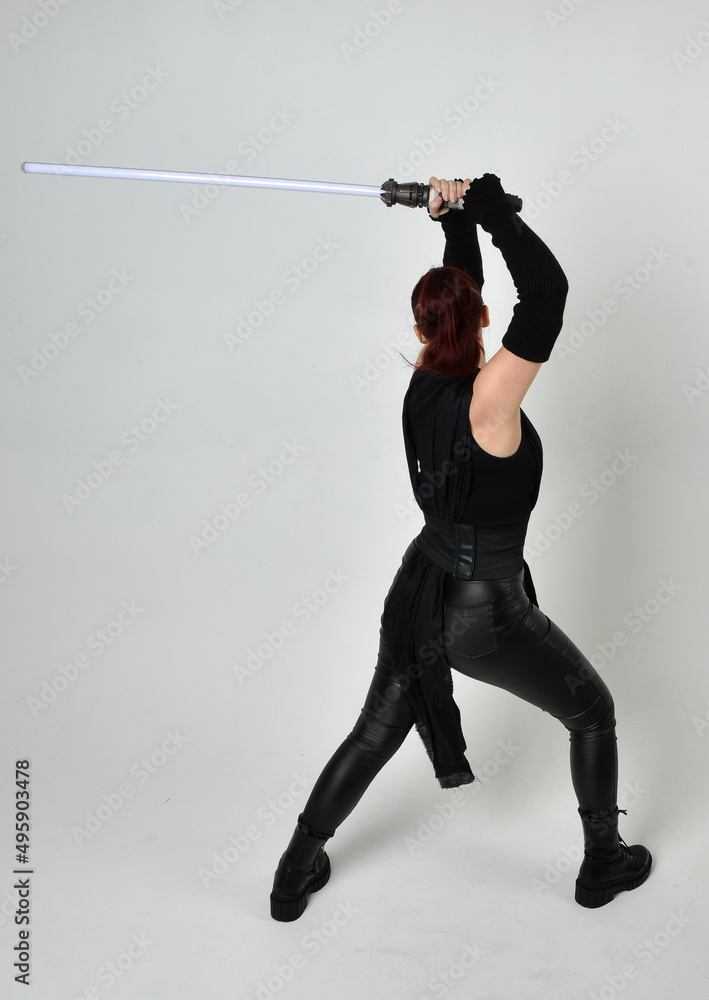 Full length portrait of pretty red haired female model wearing black futuristic scifi leather costume, holding a lightsaber sword weapon. Dynamic standing poses on a white studio background. Full length portrait of pretty red haired female model wearing black futuristic scifi leather costume, holding a lightsaber sword weapon. Dynamic standing poses on a white studio background.