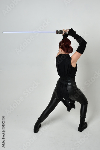 Full length portrait of pretty red haired female model wearing black futuristic scifi leather costume, holding a lightsaber sword weapon. Dynamic standing poses on a white studio background.