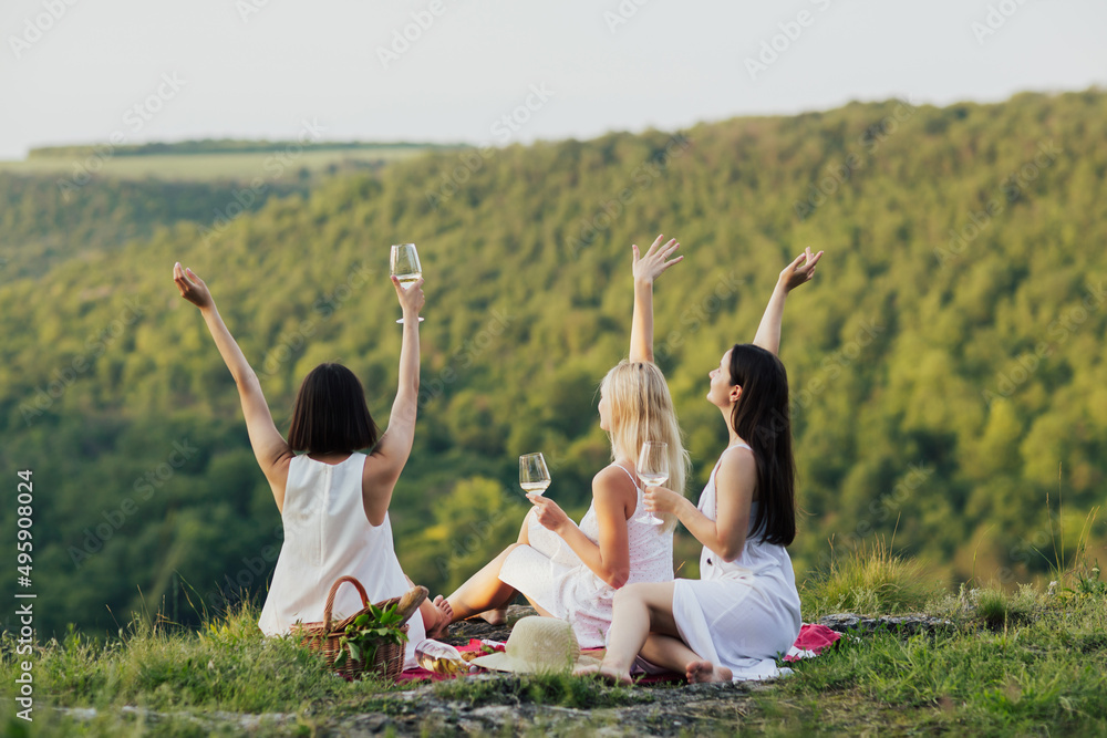 Three female friends in white dresses sitting on the grass on the hill and having fun. View from behind. Young girlfriends having a picnic with hands up.
