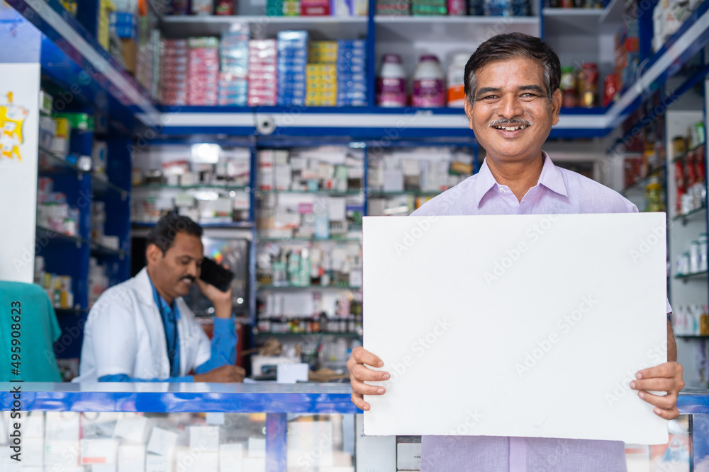 happy customer sytanding in front of medical shop by holding white empty signs board by looking at camera - concept of promotion, advertisement, promotion and happy customer.