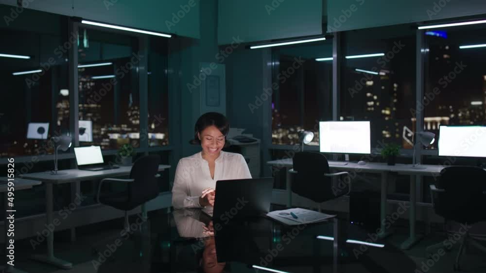 Happy African-American businesswoman work on laptop and laugh sitting at desk late in dark office Happy African-American businesswoman work on laptop and laugh sitting at desk late in dark office