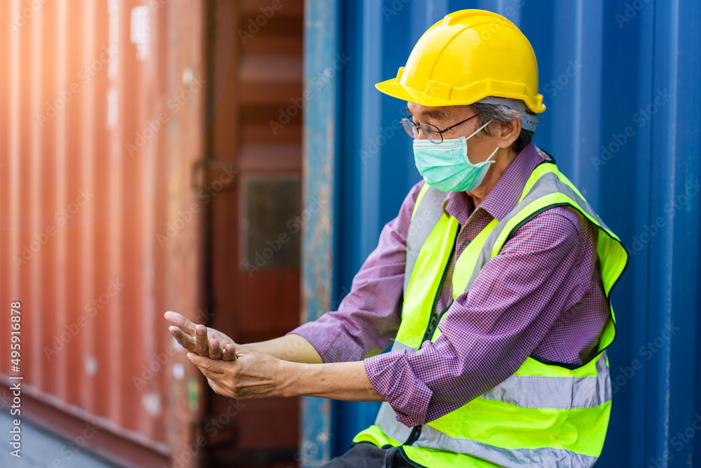 senior engineer wearing a uniform and a helmet, an engineer wearing a mask, feels pain in his fingers, stretches his fingers to relieve pain in the container courtyard. senior engineer wearing a uniform and a helmet, an engineer wearing a mask, feels pain in his fingers, stretches his fingers to relieve pain in the container courtyard.