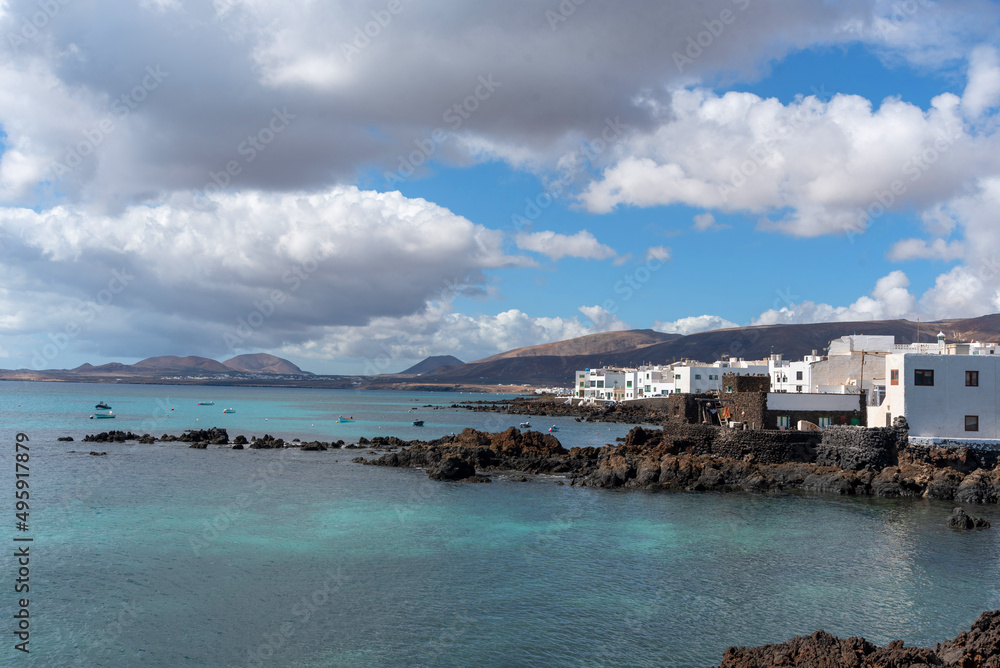 Panorámica de Punta Mujeres en Lanzarote, con casas típicas de color blanco frente a un mar cristalino y una gran montaña volcánica detrás un día nuboso, Islas Canarias.