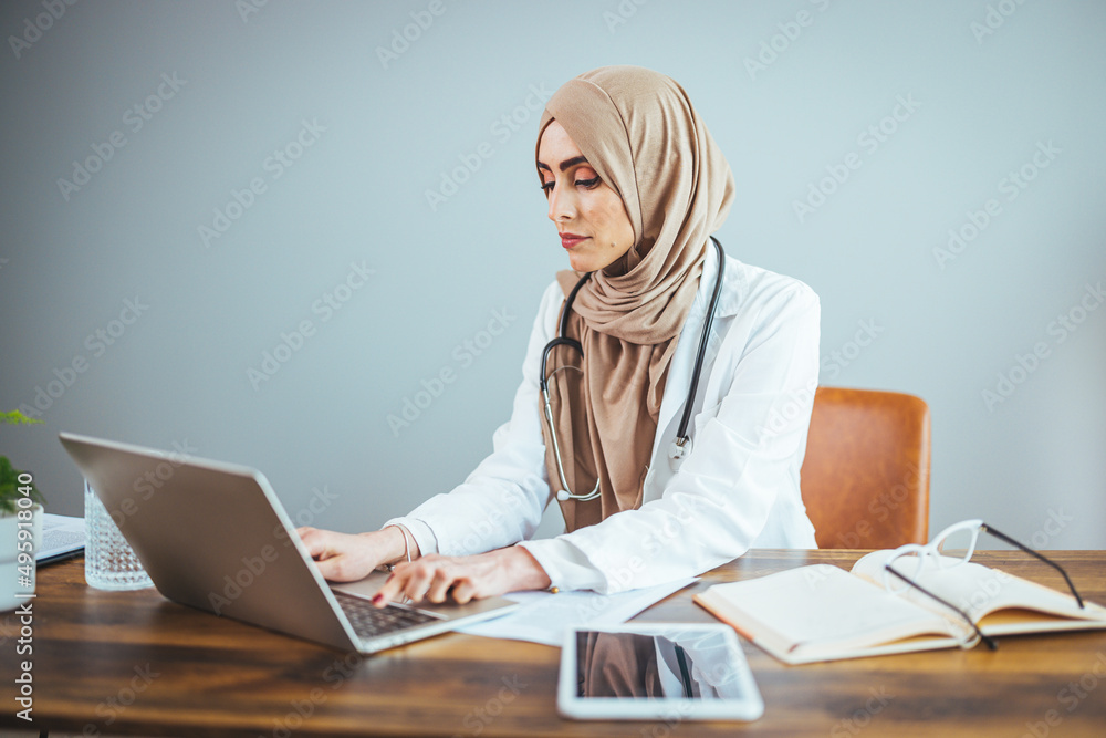 A Muslim female doctor is sitting at the table at home and working on her laptop. A happy and confident Muslim woman is using her laptop in her living room. A Muslim female doctor is sitting at the table at home and working on her laptop. A happy and confident Muslim woman is using her laptop in her living room.