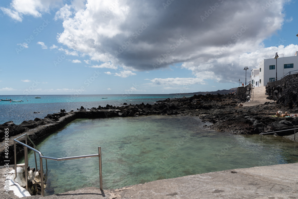 Piscina natural de agua turquesa y transparente junto a una casa típica de color blanca en Punta mujeres, Lanzarote durante un día soleado con una gran nube. Recursos naturales de las Islas Canarias.