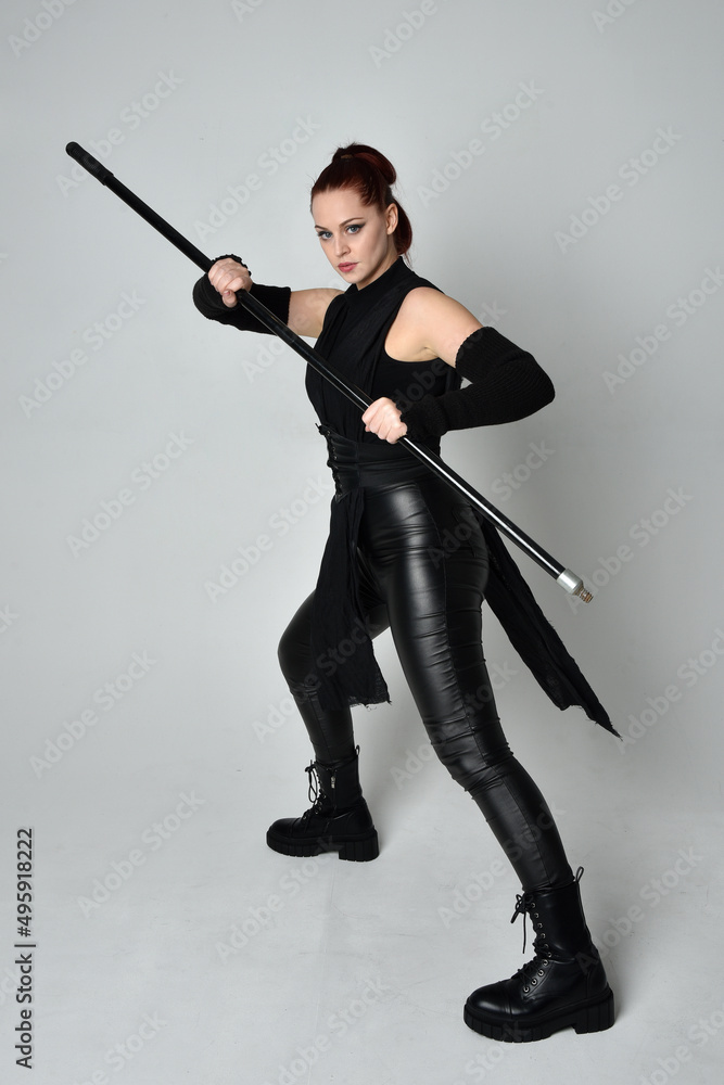 Full length portrait of pretty red haired female model wearing black futuristic scifi leather costume, holding a staff spear weapon. Dynamic standing pose on a white studio background.