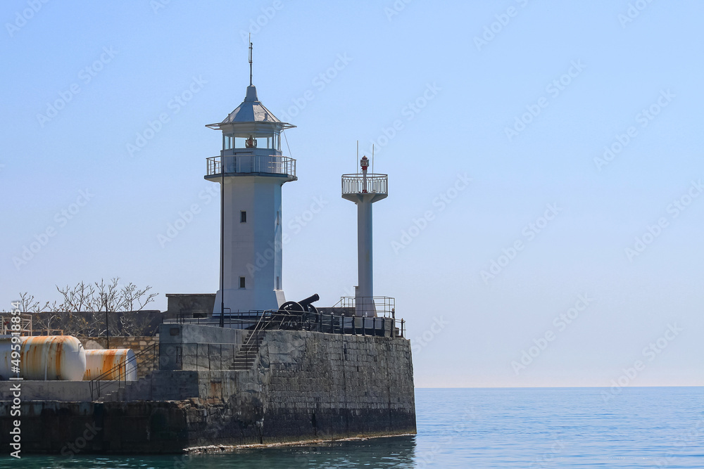 The white tower of a small old lighthouse on a rocky promontory. In the background is a calm blue sea and sky. Yalta, Crimea.