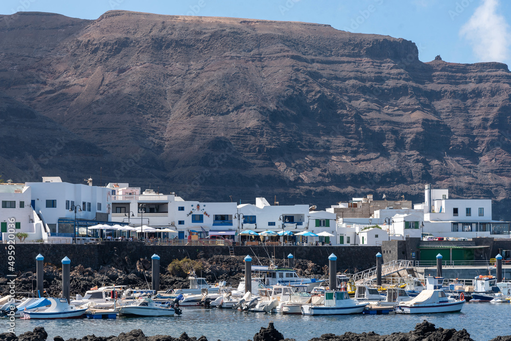 Pequeño pueblo de casas blancas tradicionales de Lanzarote, frente a un pequeño puerto lleno de barcos y con una gran montaña al fondo durante un día soleado. Recursos turísticos de Islas Canarias.