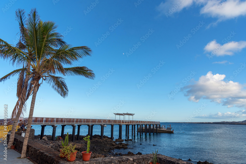 Vista paradisiaca de Lanzarote Islas Canarias, un muelle adentrándose en el mar turquesa con una gran palmera al lado durante un día soleado con el cielo azul despejado. Recursos naturales de Canarias