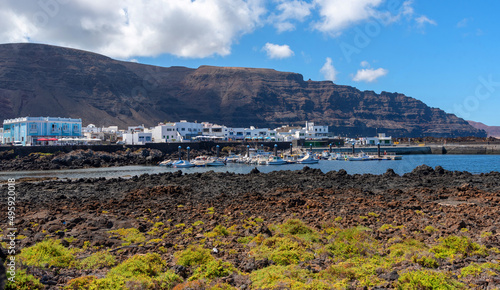 Pueblo de la costa de Lanzarote con una gran montaña al fondo lleno de casas tradicionales blancas con un puerto delante con marea baja un día de verano soleado. Recursos naturales de Islas Canarias. photo