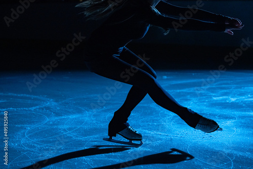 Figure skating lady is wearing black sportswear is skating on ice rink, training at night in the rays of blue light. Young woman preparing to competition, training rotation and slides skills. Close up