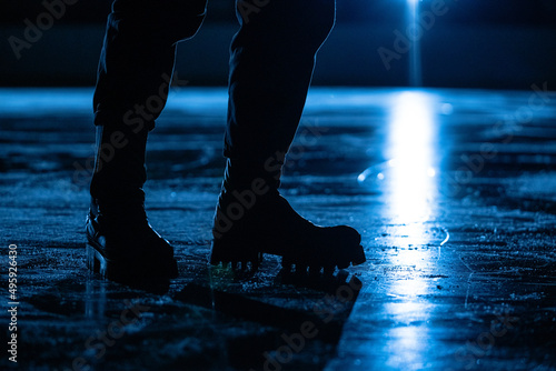 Men's legs in rough boots on the ice of a sports arena. A male coach stands on surface of an ice stadium for figure skating. A beam of blue spotlight reflects off shiny, scratched ice. Close up.