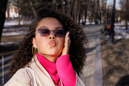 Happy curly brunette girl in beige hat and sun glasses smiling outdoors. Young woman happy walking in street. Pink sweater, beige coat, beige hat. Sun in city. Fashionable asian girl with frizzly hair © Tatyana