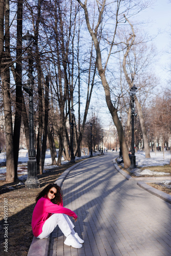 Happy curly brunette girl in sun glasses and hat smiling outdoors. Young woman happy walking in street. Pink sweater, beige coat, beige hat. Sun in city. Fashionable asian girl with frizzly hair