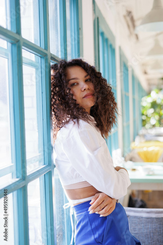 Serious curly girl in cafe, thoughful. Woman in white blouse and blue trousers closed and distrustful in sun light Free Download Serious curly girl in cafe, thoughful. Woman in white blouse and blue trousers closed and distrustful in sun light - GFXLoad