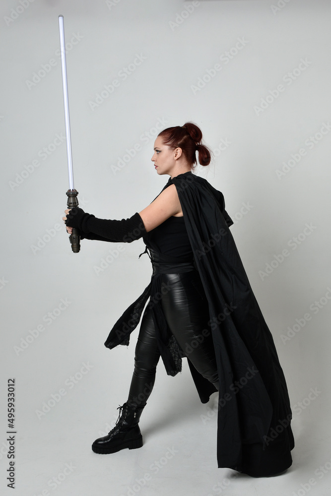 Full length portrait of pretty redhead female model wearing black futuristic scifi leather cloak costume, holding a lightsaber weapon. Dynamic standing pose on white studio background. Full length portrait of pretty redhead female model wearing black futuristic scifi leather cloak costume, holding a lightsaber weapon. Dynamic standing pose on white studio background.