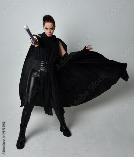 Full length portrait of pretty redhead female model wearing black futuristic scifi leather cloak costume, holding a lightsaber weapon. Dynamic standing pose on white studio background.