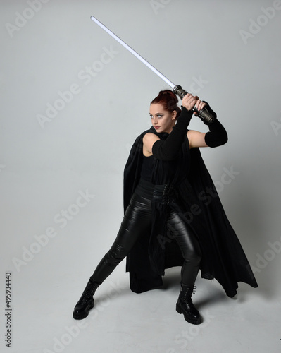 Fotografie Full length portrait of pretty redhead female model wearing black futuristic scifi leather cloak costume, holding a lightsaber weapon