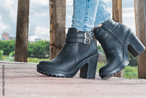 close-up detail of a latina girl in blue jeans and black synthetic leather ankle boots posing with her feet crossed, standing on a wooden balcony with brown railings. fashion concept photo