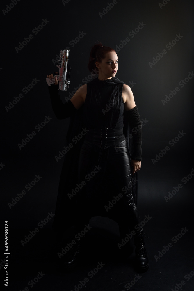 Full length portrait of pretty redhead female model wearing black futuristic scifi leather cloak costume, holding a gun weapon. Dynamic standing pose on studio background. Full length portrait of pretty redhead female model wearing black futuristic scifi leather cloak costume, holding a gun weapon. Dynamic standing pose on studio background.