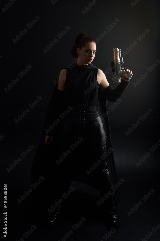 Full length portrait of pretty redhead female model wearing black futuristic scifi leather cloak costume, holding a  gun weapon. Dynamic standing pose on  studio background.