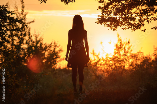 Fotografie Back view of young woman in summer dress walking alone through evening dark fore