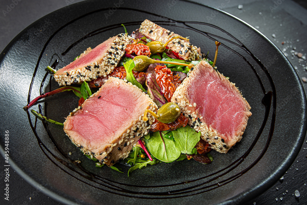 Close up of tuna slices withsesam seeds and fresh vegetables on a dark plate.