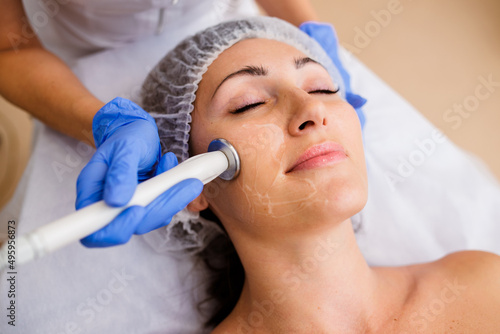 Gorgeous woman with smooth skin getting cosmetology procedures in modern beauty salon. Hands of cosmetology specialist holding device and doing radio frequency facial treatment, softening wrinkles.